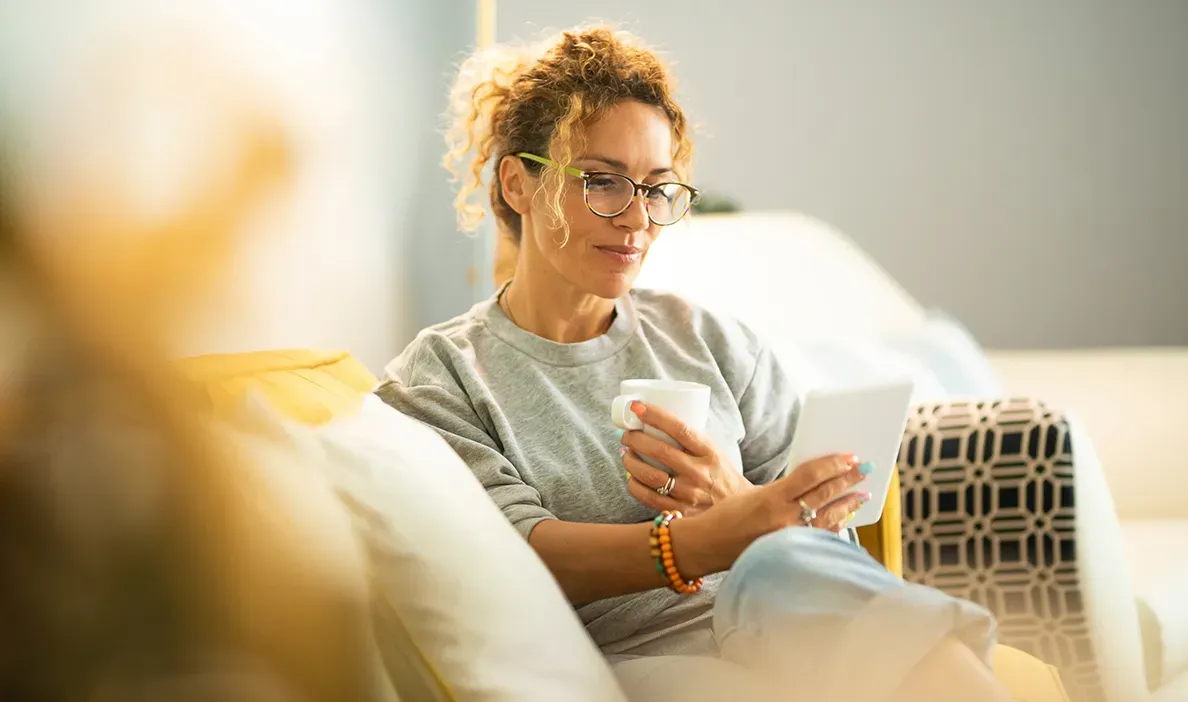 Woman with curly blonde hair, wearing glasses, sitting on a cozy couch while sipping coffee and reading on her phone.