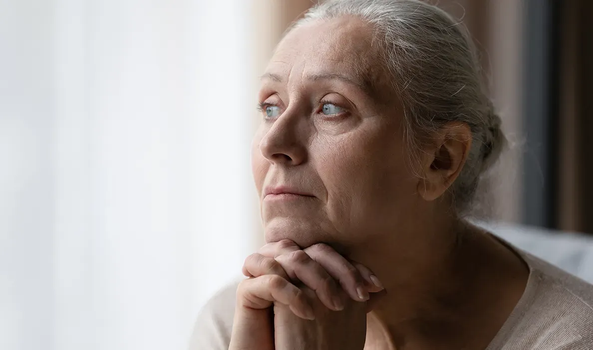 Woman with grey hair gazing into the distance with hands clasped under her chin, appearing deep in thought.