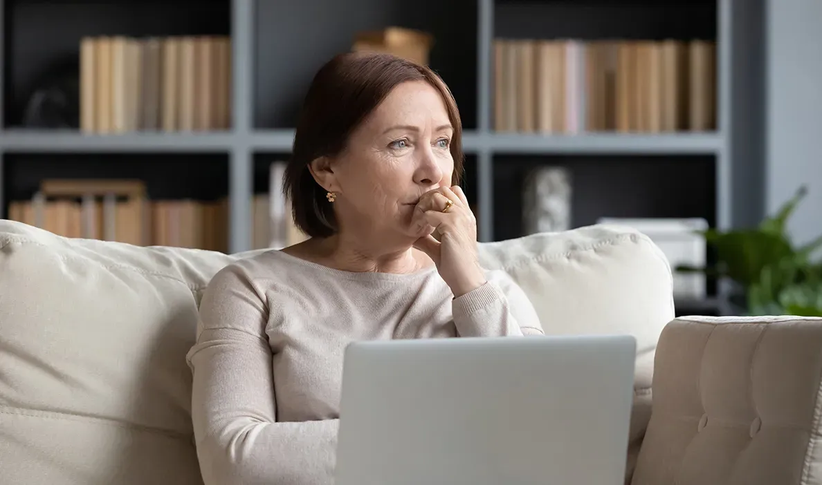 Woman sitting on a couch with a laptop on her lap, looking deep in thought while biting her finger.
