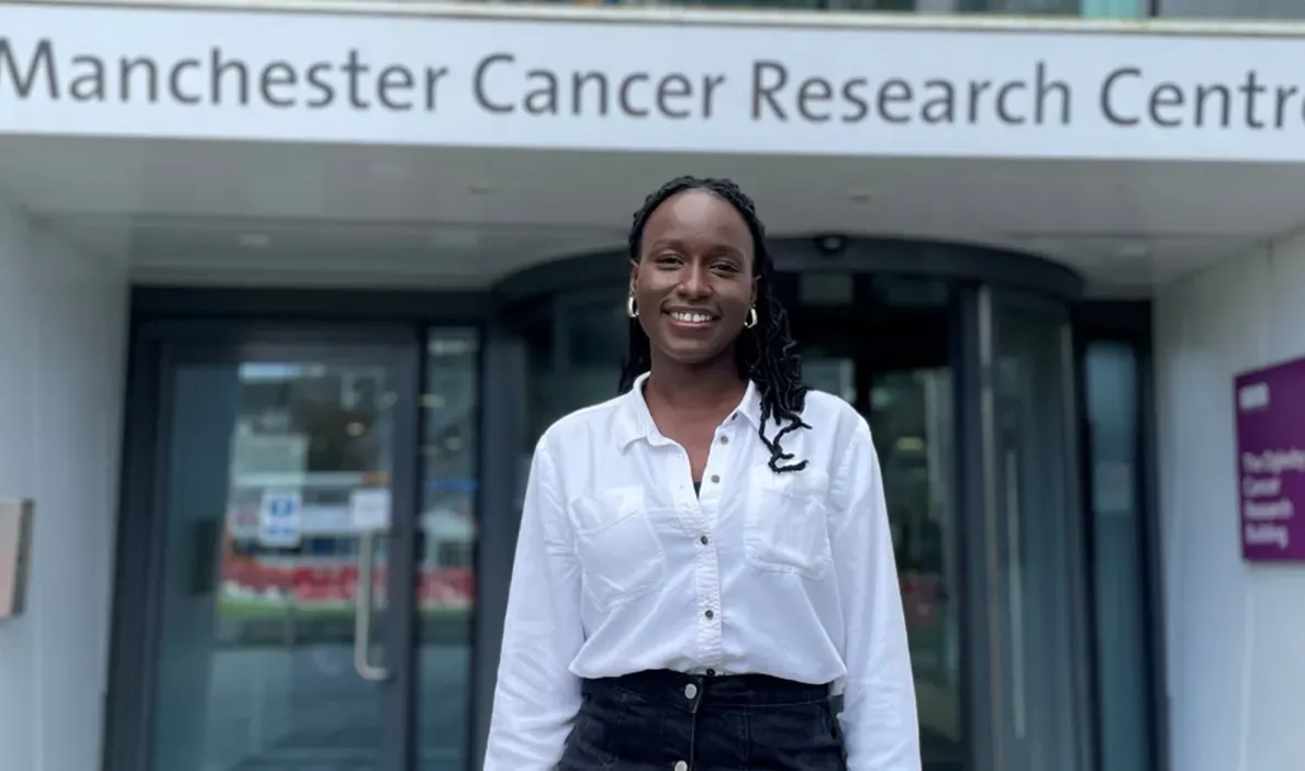 Woman smiling outside the Manchester Cancer Research Centre, wearing a white shirt and standing in front of the entrance.