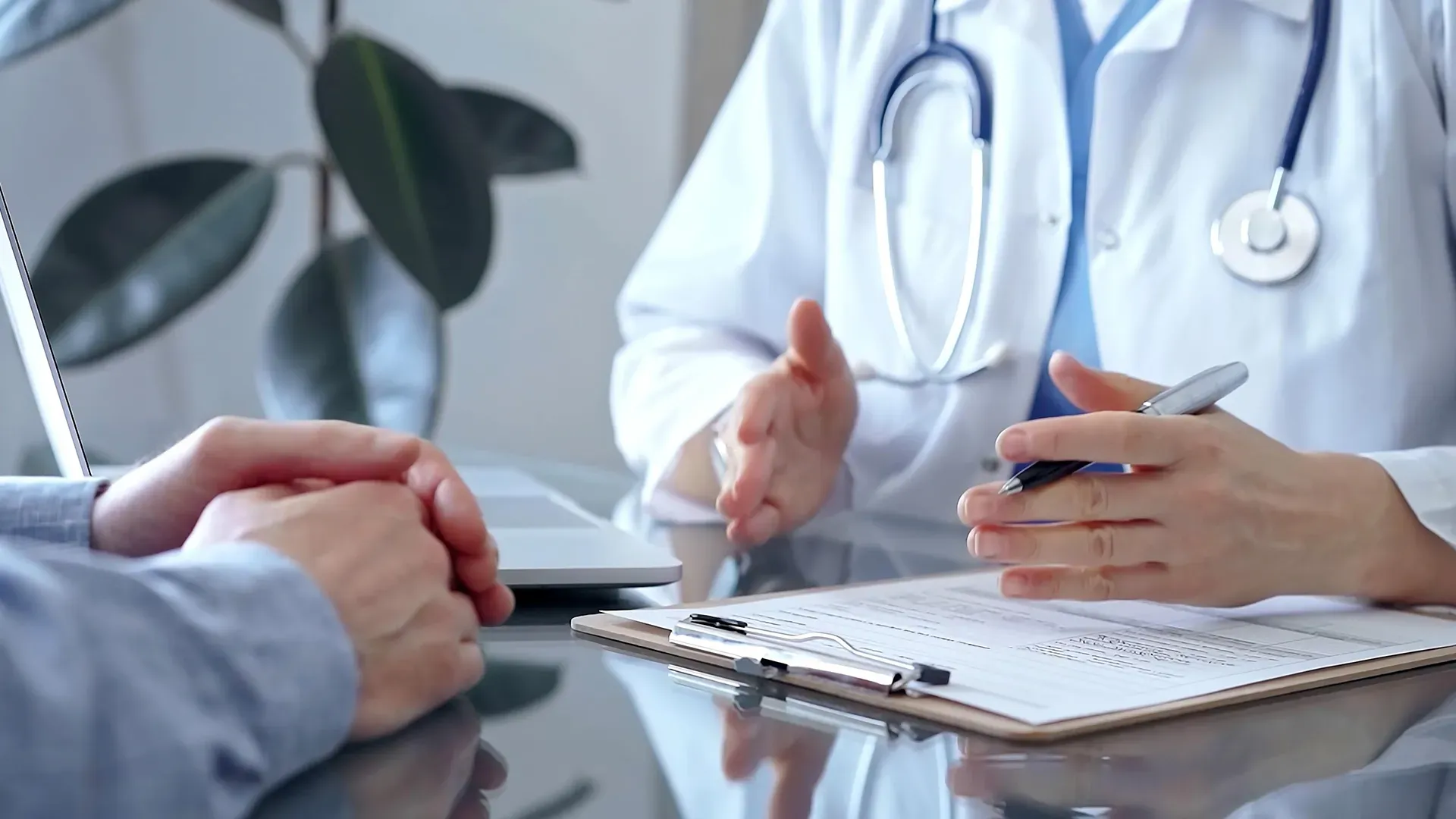 Doctor in a white coat with a stethoscope discussing test results with a patient. The doctor's hands are gesturing while holding a pen.