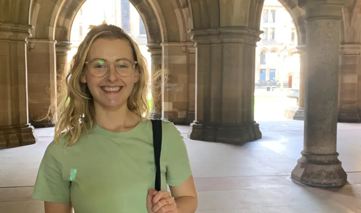 Woman smiling under stone archways at a historic university building, wearing glasses and a green T-shirt.