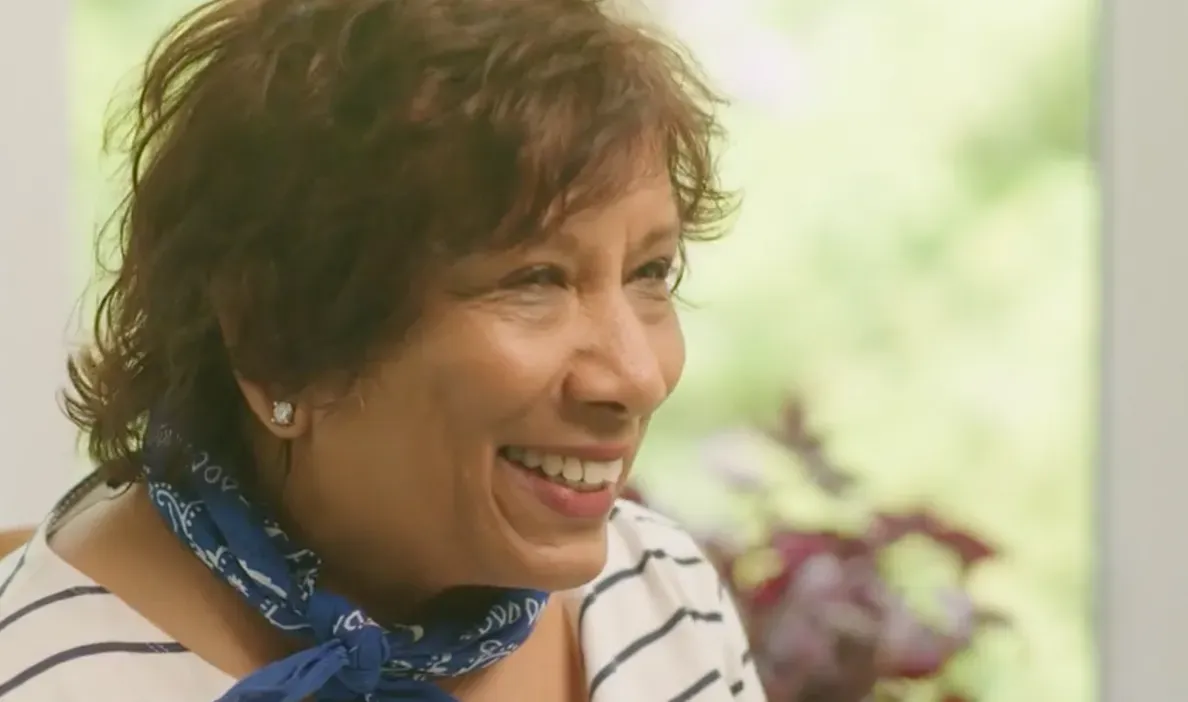 A smiling woman with short, wavy brown hair is wearing a striped shirt and a blue bandana around her neck.